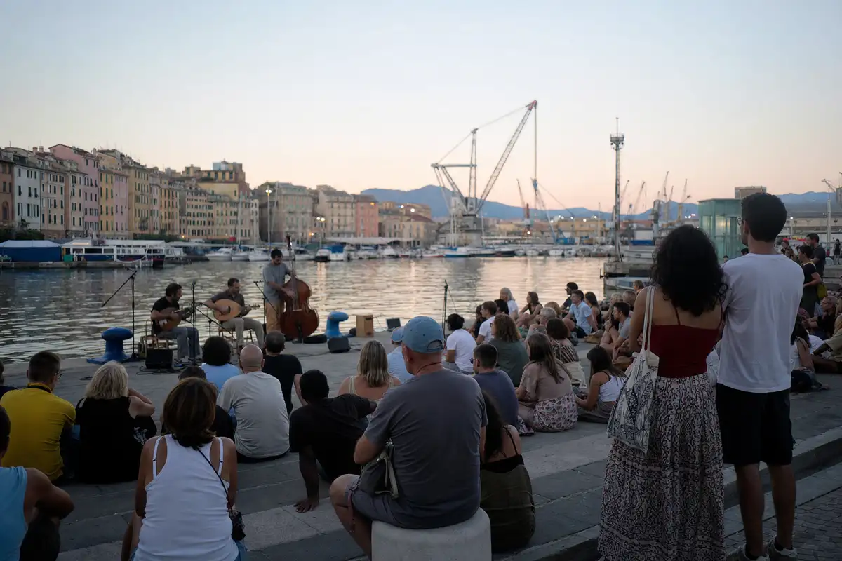 Pubblico seduto sul molo ascolta un trio acustico al porto, con barche e gru sullo sfondo all’alba