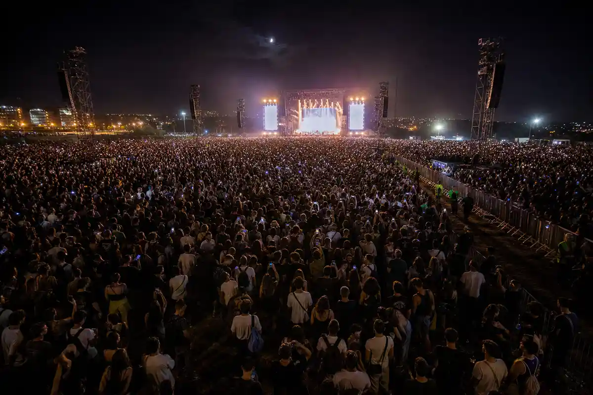 Folla enorme a un concerto serale all’aperto, con palco illuminato in lontananza e smartphone alzati