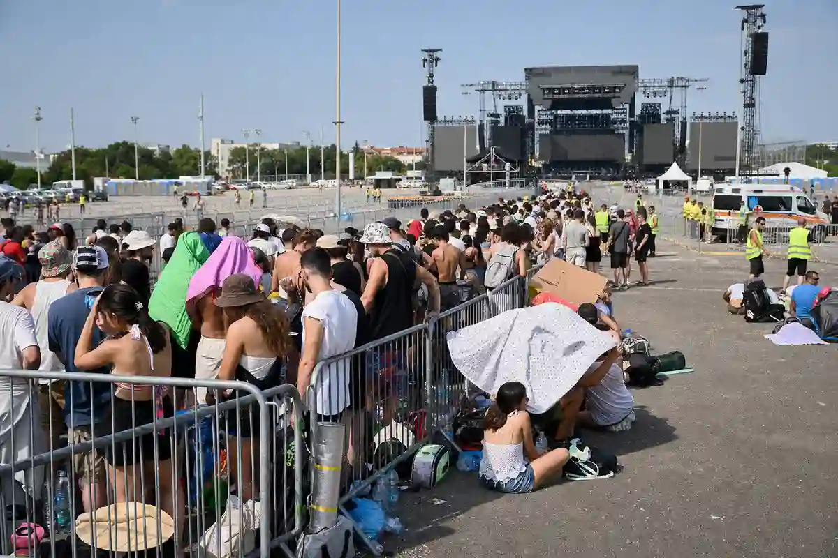 Fan in fila dietro transenne sotto il sole, con zaini e bottiglie d’acqua, e grande palco in lontananza