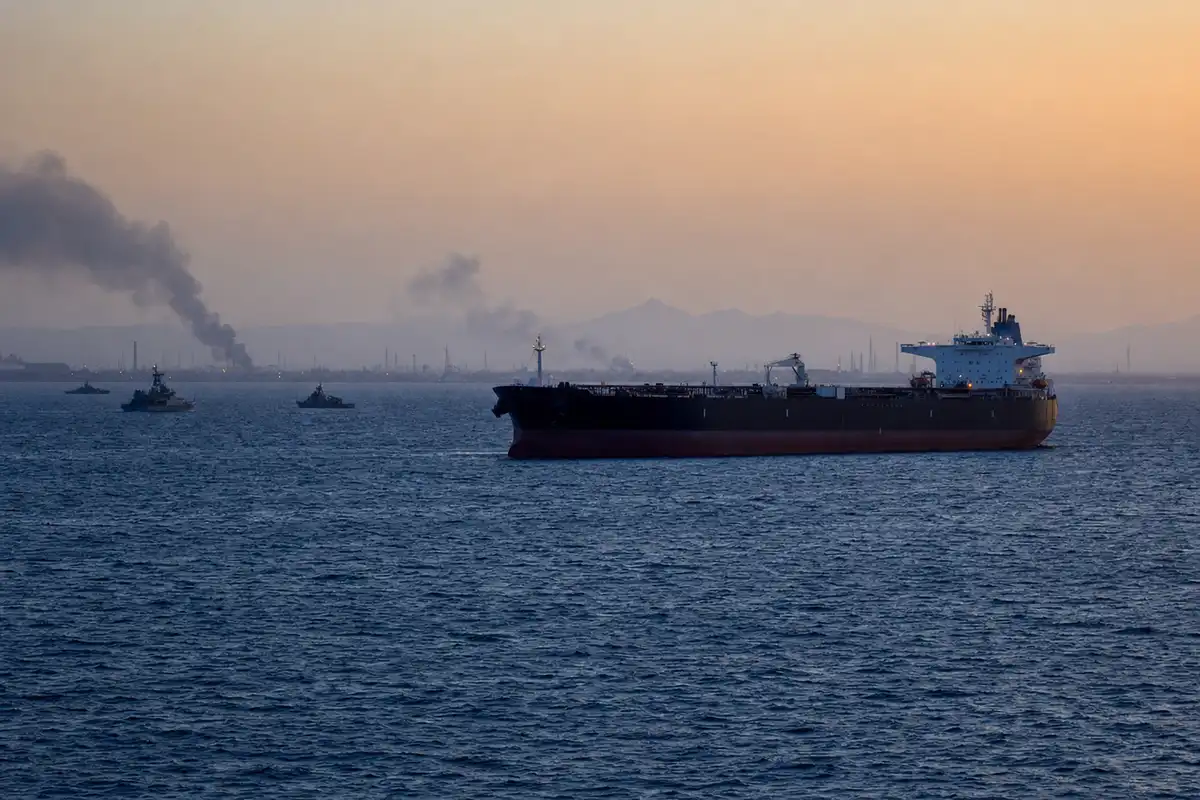 Petroliera in mare al tramonto con piccole navi militari sullo sfondo e fumo all’orizzonte